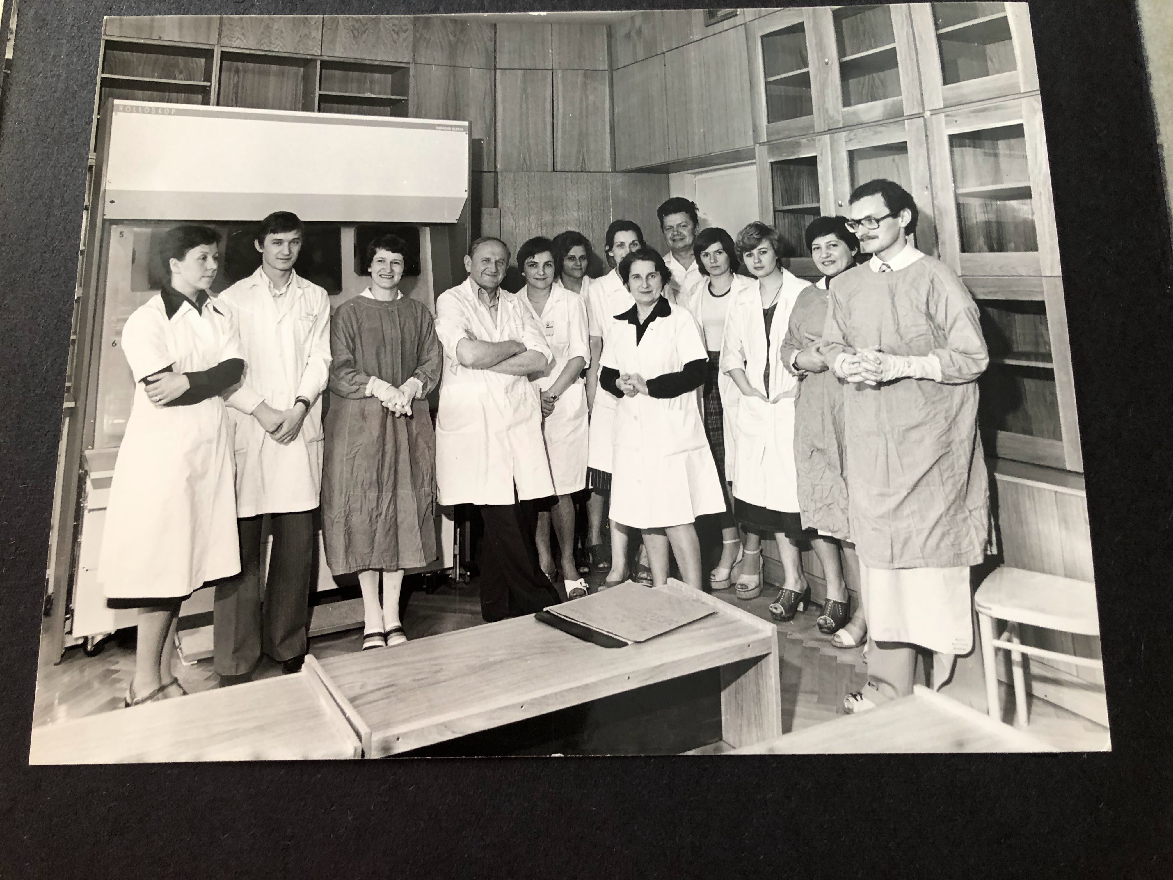 Czarno-biała fotografia grupowa przedstawiająca kilkunastoosobowy zesp&oacute;ł lekarzy i technik&oacute;w w fartuchach ochronnych i białych kitlach, stojących razem w pomieszczeniu pracowni radiologicznej.